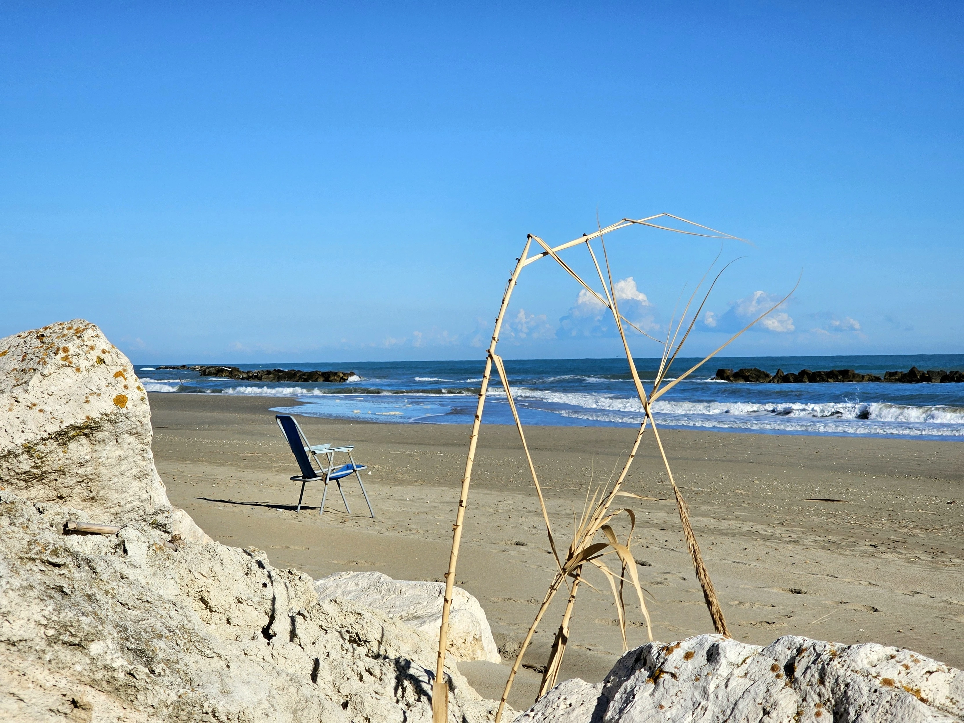 A tranquil beach scene with a single blue folding chair set up on the sand near the water's edge. In the foreground, a large rock and a few sparse strands of beach grass are visible. The ocean is calm with small waves breaking over a line of rocks in the distance, under a clear blue sky with a few scattered clouds.