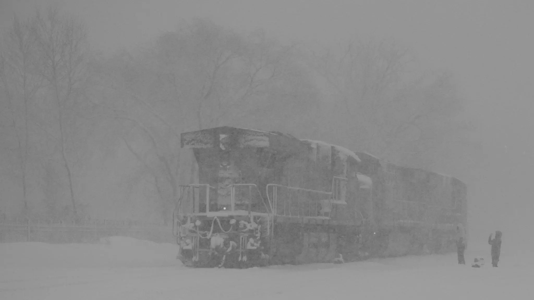 A pair of diesel locomotives moving slowly through a near-whiteout blizzard. Two children wave to it. 