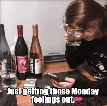 A girl sits at a table, the table has multiple bottles of alcohol on it.  There are multiple packs of cigarettes open, the girl is smoking a cigarette and drinking from a glass while looking at their phone.  They appear to be very distressed.  There is text on this image that indicates "Just getting those Monday feelings out."