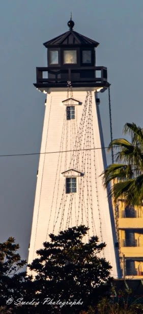 "The image shows a lighthouse in Gulfport, Mississippi. The lighthouse is white and has a tapered structure with a black lantern room at the top. The lantern room features windows on all sides and is topped with a black, pointed roof. Strings of lights are draped from the top of the lighthouse down to its base, suggesting it may be decorated for a festive occasion. In the foreground, there are trees and a palm tree, and part of a yellow building is visible to the right of the lighthouse. The sky in the background is clear, indicating a sunny day. The image is signed "© Swede's Photographs" at the bottom." - Copilot