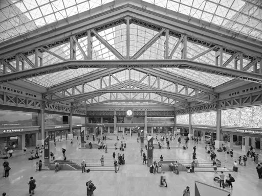 A modern urban passenger rail station hall. Passengers mill about around escalators that descend to unseen tracks below. Exposed steel beams support a skylight roof.