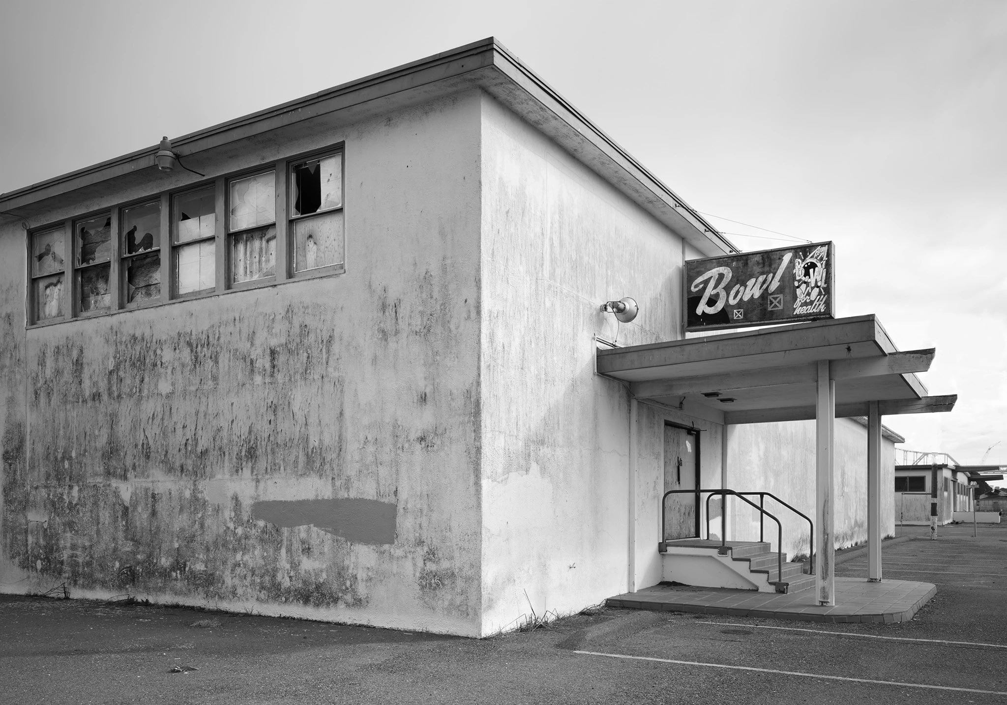 A simple industrial-type structure, seen from the corner. A row of broken windows on the top of one side. Doorway with awning and sign reading "Bowl for Health", with bowling icons, on other side.