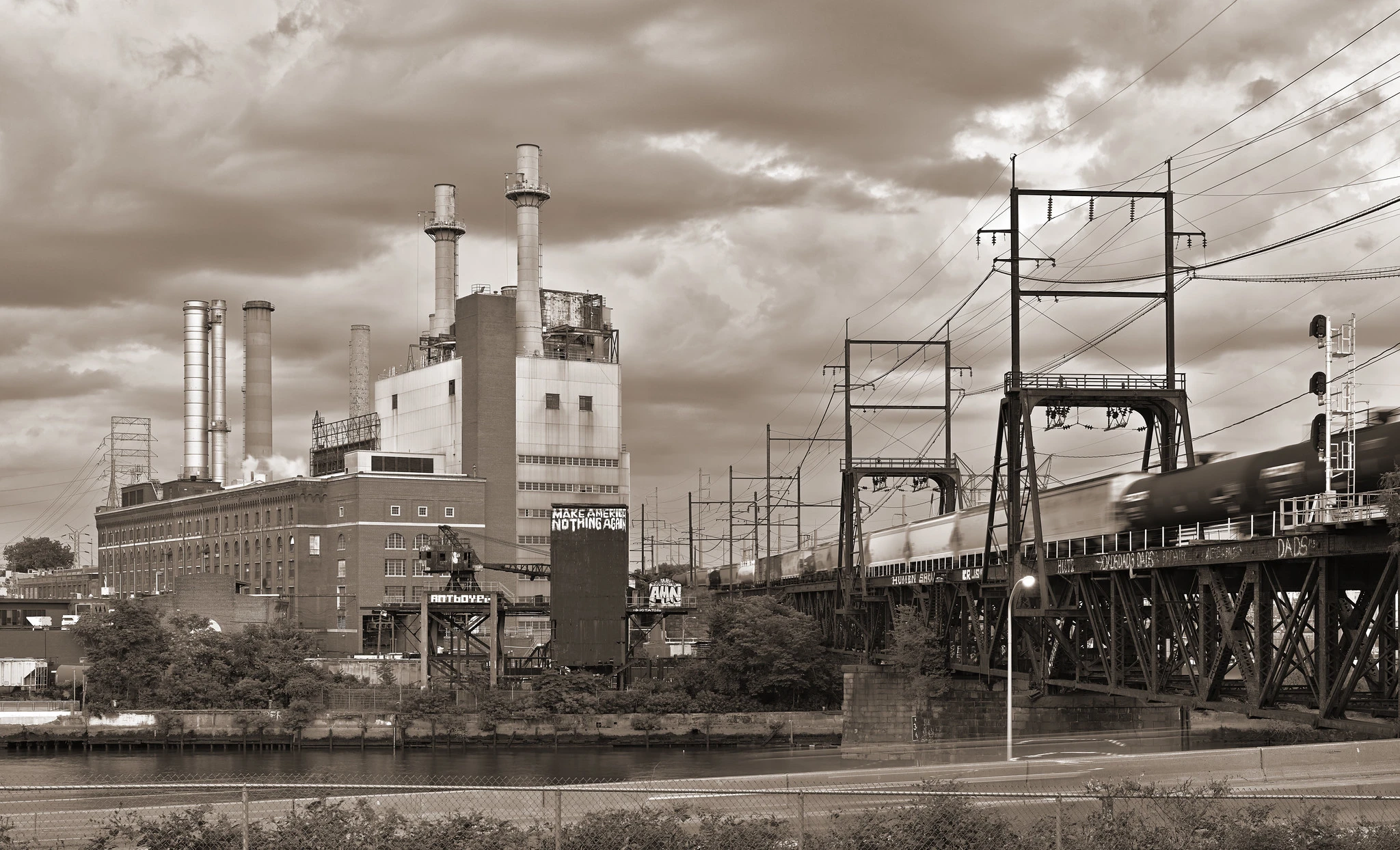 A mid-century power plant, with four prominent smokestacks, at left across a small river. At right, a railroad drawbridge crosses the river, with a freight train streaking across.