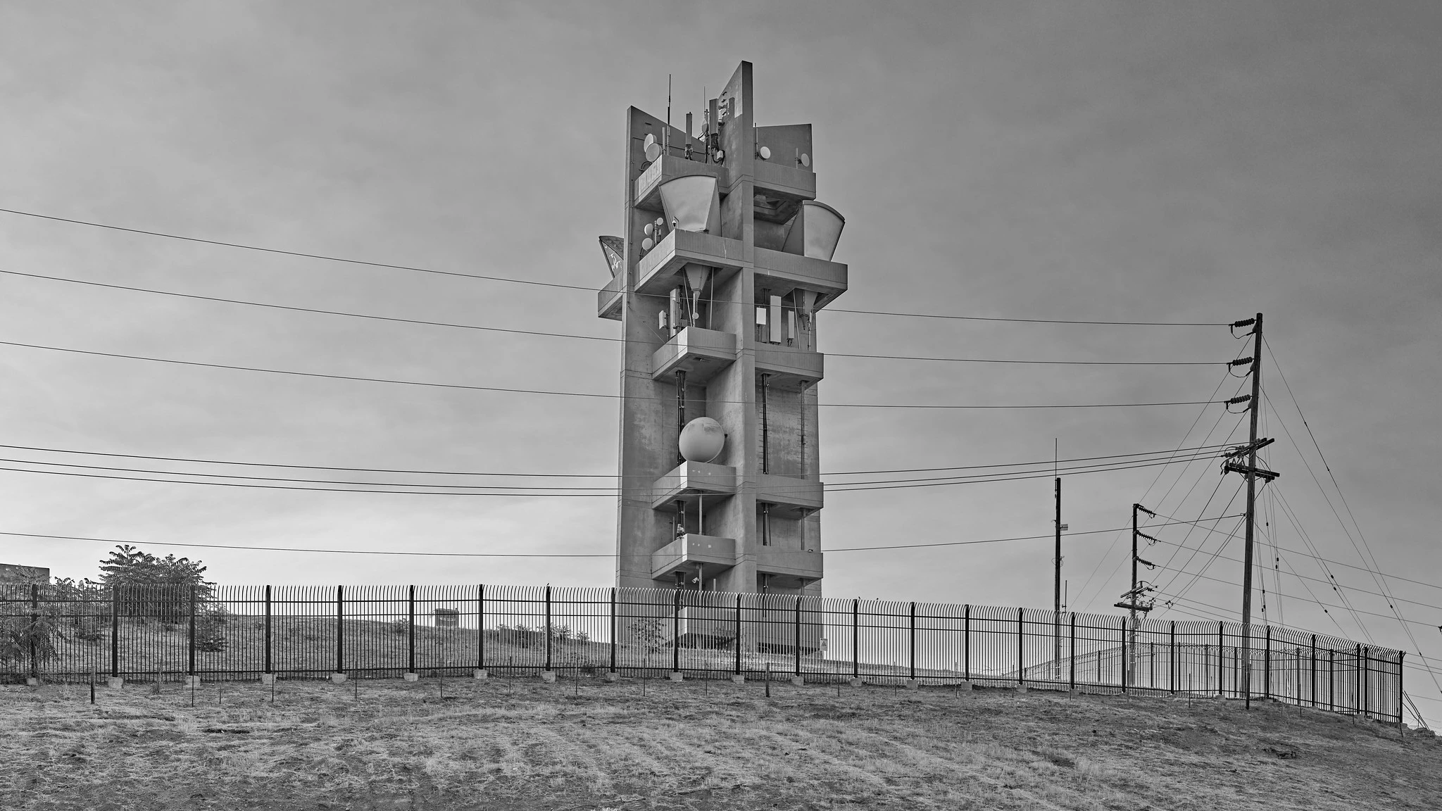 A heavy-duty concrete tower, with multiple levels of platforms supporting microwave antennas, including "horn" antennas, on a hilltop behind a fence.