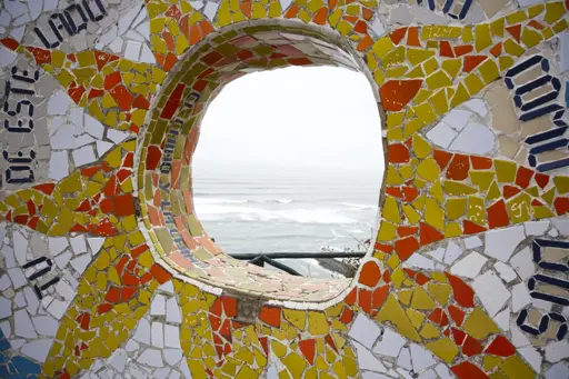 A gray ocean under gray skies can be seen in a window in a colorful yellow and red flower petal mosaic. Parque del Amor, Lima, November 2024. Photo by Peachfront.