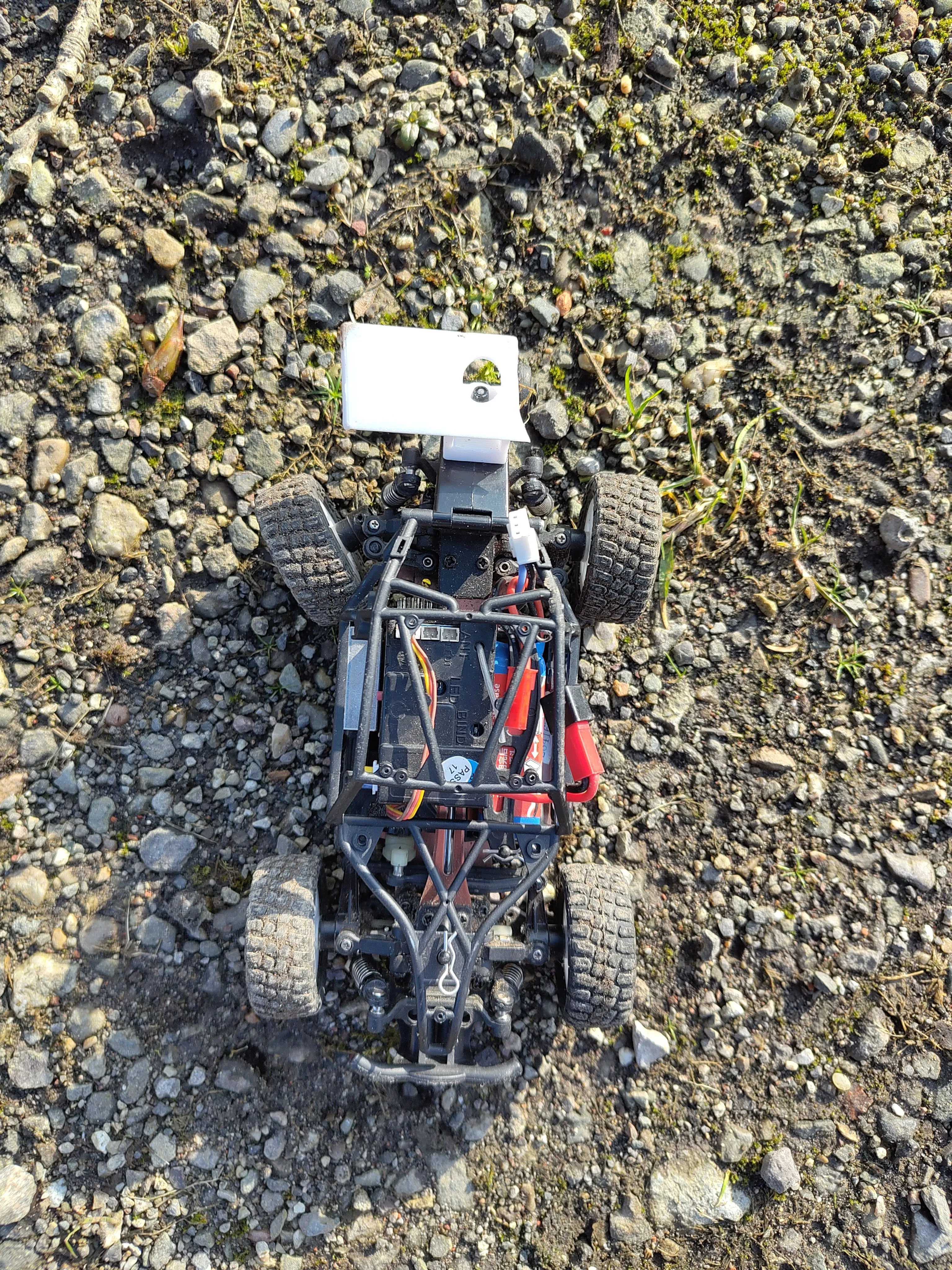 a top-down photo of a WLLtoys 284161 RC car with a broken spoiler and a broken rear wheel connection, it is sitting on gravel