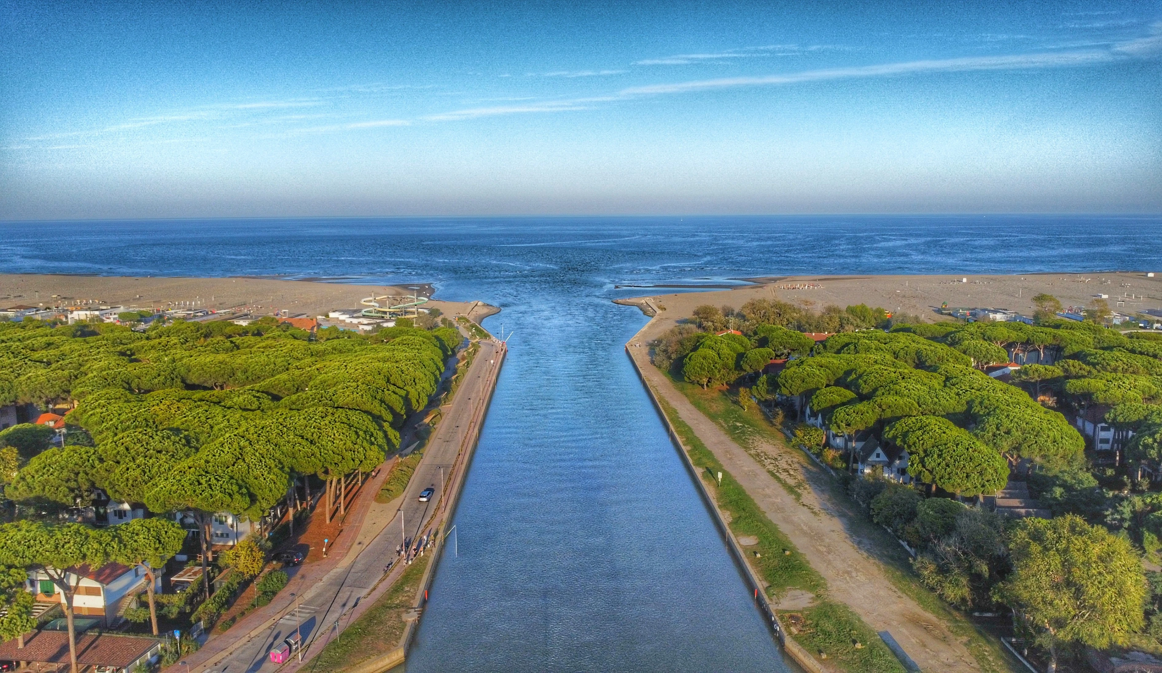 Aerial view of a serene landscape featuring a long, narrow waterway leading to a vast, blue sea. On either side of the waterway are lush green trees, resembling a canopy, with houses nestled beneath. A road runs parallel to the water, with a few pedestrians visible. Beyond the trees, there's a sandy beach with some people and amenities, and further out, the expansive sea stretches to the horizon. The sky is clear with a slight gradient from blue to a lighter shade near the horizon.