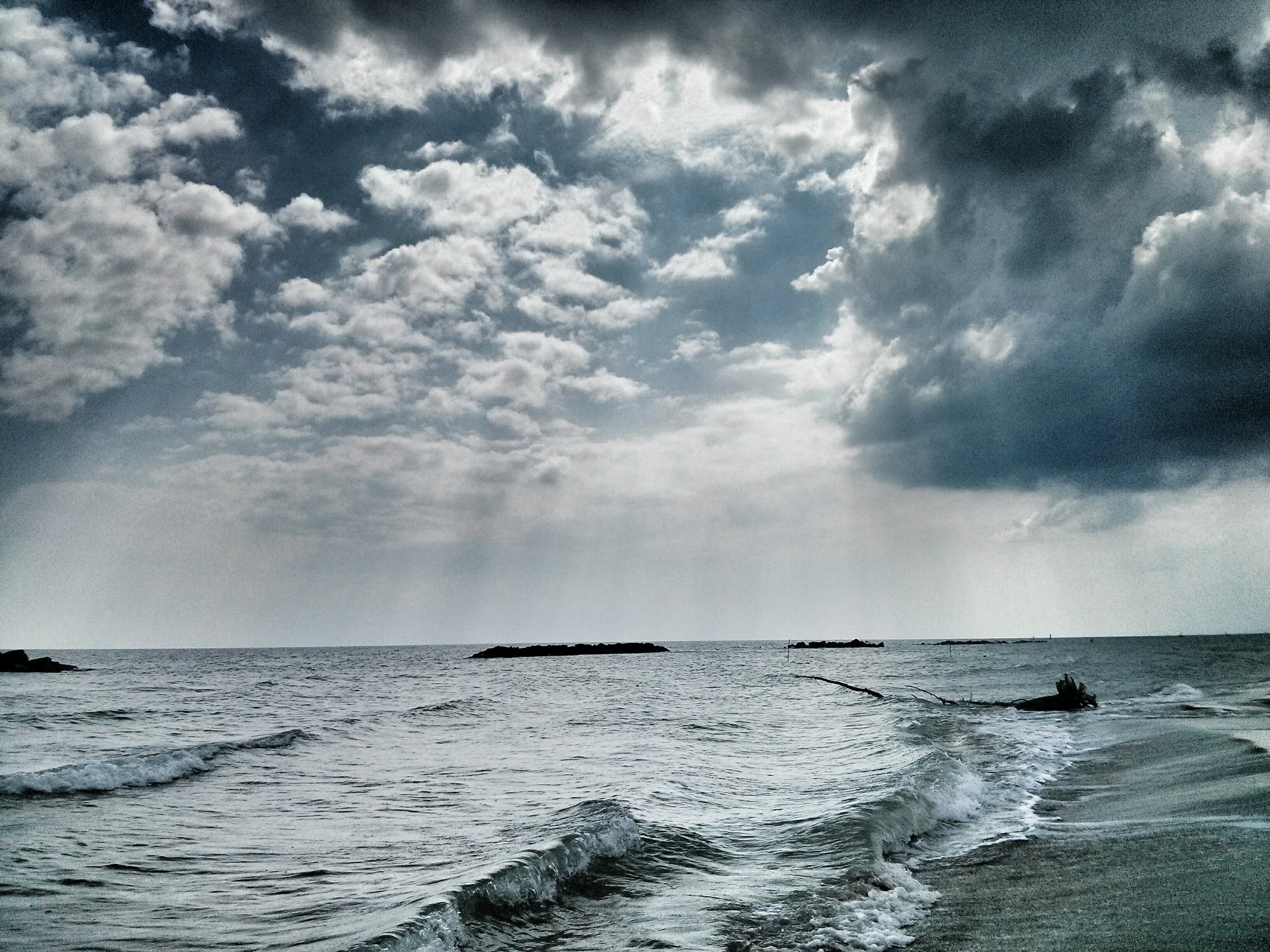 A peaceful seaside view under a dramatic, overcast sky, with a mix of heavy clouds and sunlight streaming through gaps. The waves gently lap at the shore, creating a soft, rhythmic sound that contrasts with the looming clouds. Despite the strength of nature's presence, the scene evokes a perfect sense of silent solitude, as if the power of the elements enhances the serenity of this quiet Sunday moment. Driftwood floats in the distance, further emphasizing the natural flow of time and calmness.