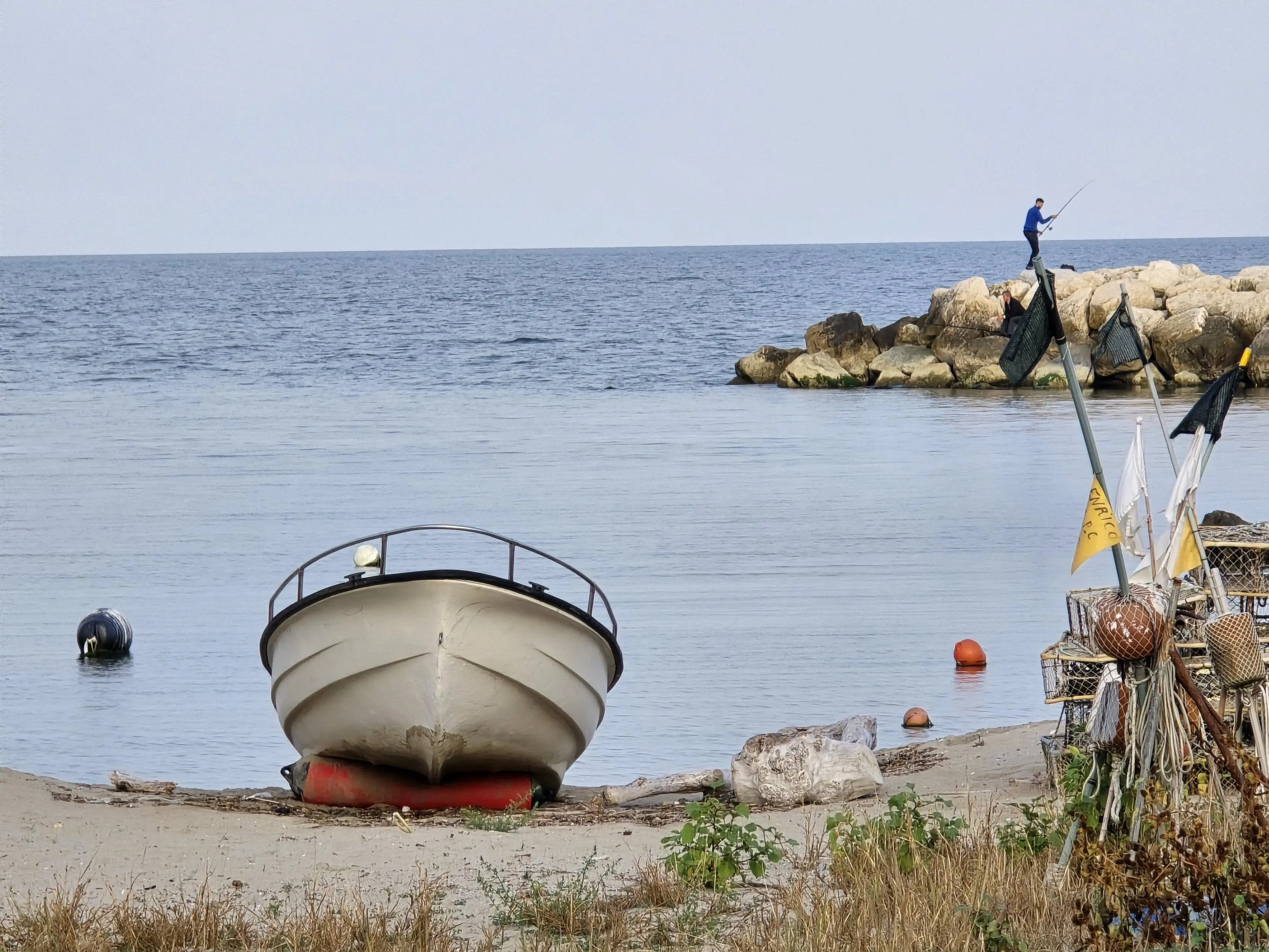 A small boat rests on a sandy shore, with fishing equipment and nets piled nearby. In the background, a lone fisherman stands on a rocky jetty, casting his line into the calm, open sea. The tranquil waters stretch out under an overcast sky, creating a peaceful, contemplative scene by the coast. Buoys and rocks add subtle colors to the muted coastal landscape, embodying the quiet rhythm of a seaside morning.