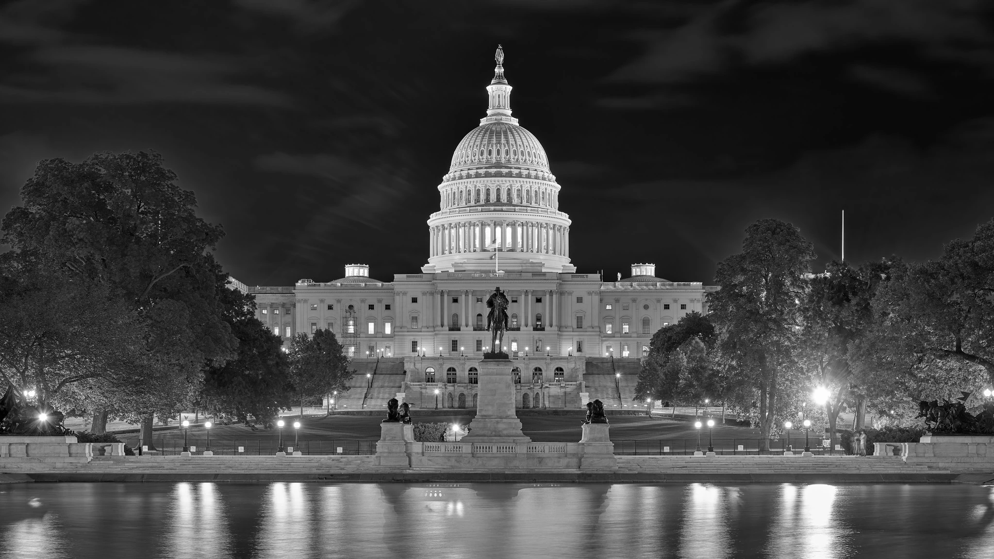 The northern side of the US Capitol building at night, with its prominent dome illuminated. Reflexions are visible in the pool at front.