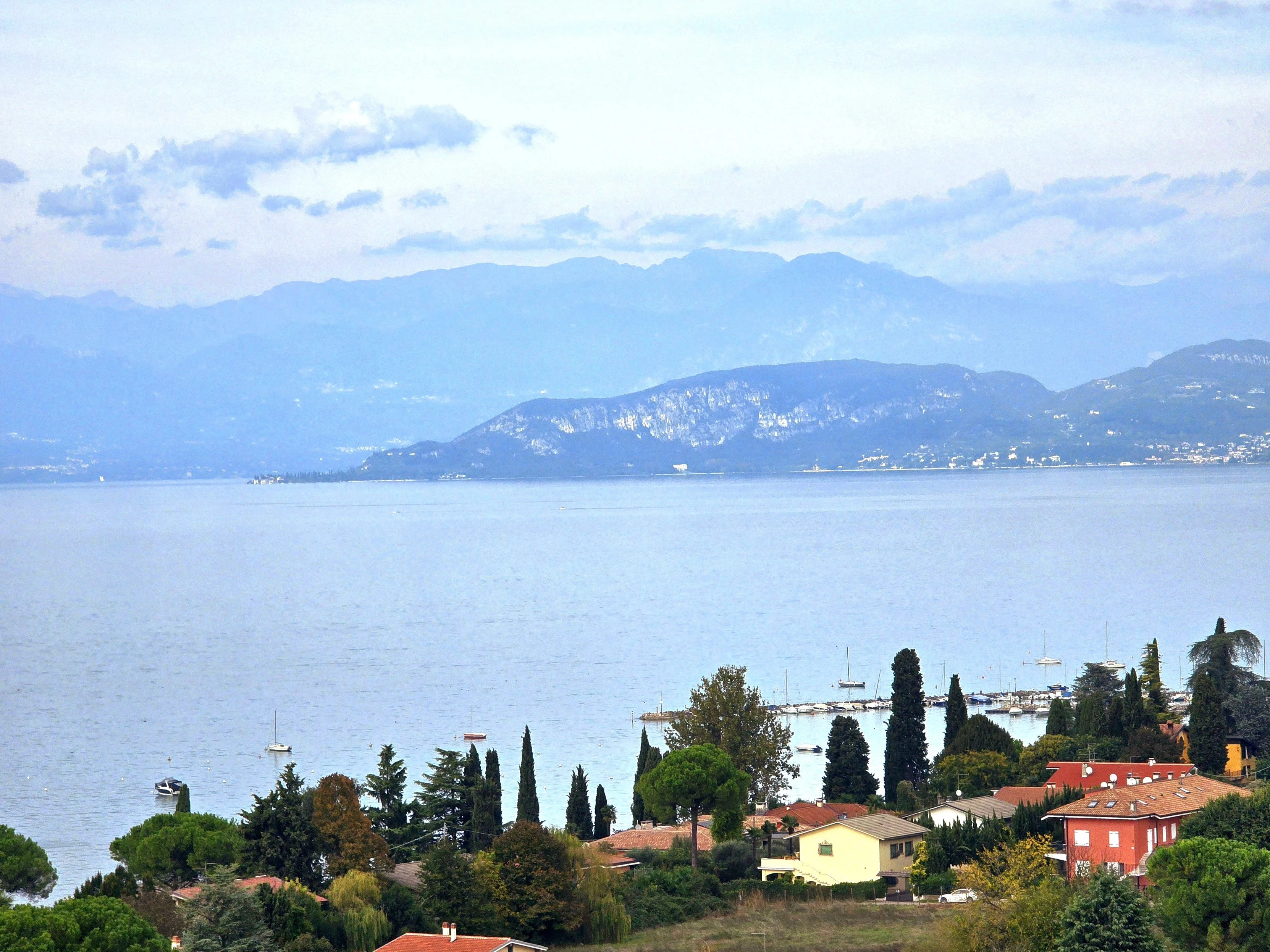 A serene view of Lake Garda in Italy, showcasing calm waters with gentle ripples reflecting the overcast sky. In the foreground, a charming lakeside village is nestled among tall cypress and pine trees, with boats docked at a small marina. The scene is framed by distant, mist-covered mountains that rise softly in the background, adding depth and tranquility to the landscape. The overall atmosphere is peaceful, evoking a sense of quiet retreat and natural beauty.