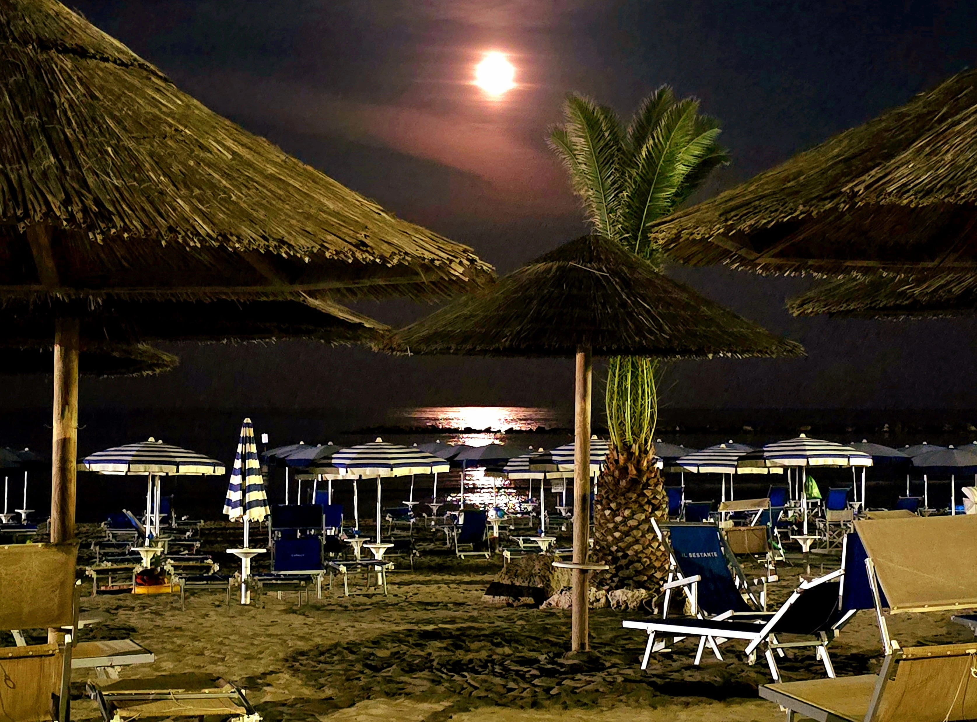 A moonlit beach at night, signaling summer's end but the sea's eternal allure. Thatched umbrellas cast long shadows over empty lounge chairs and striped parasols. A lone palm tree stands sentinel as the full moon's reflection shimmers on the dark water, reminding us that while seasons change, the sea's beauty is timeless