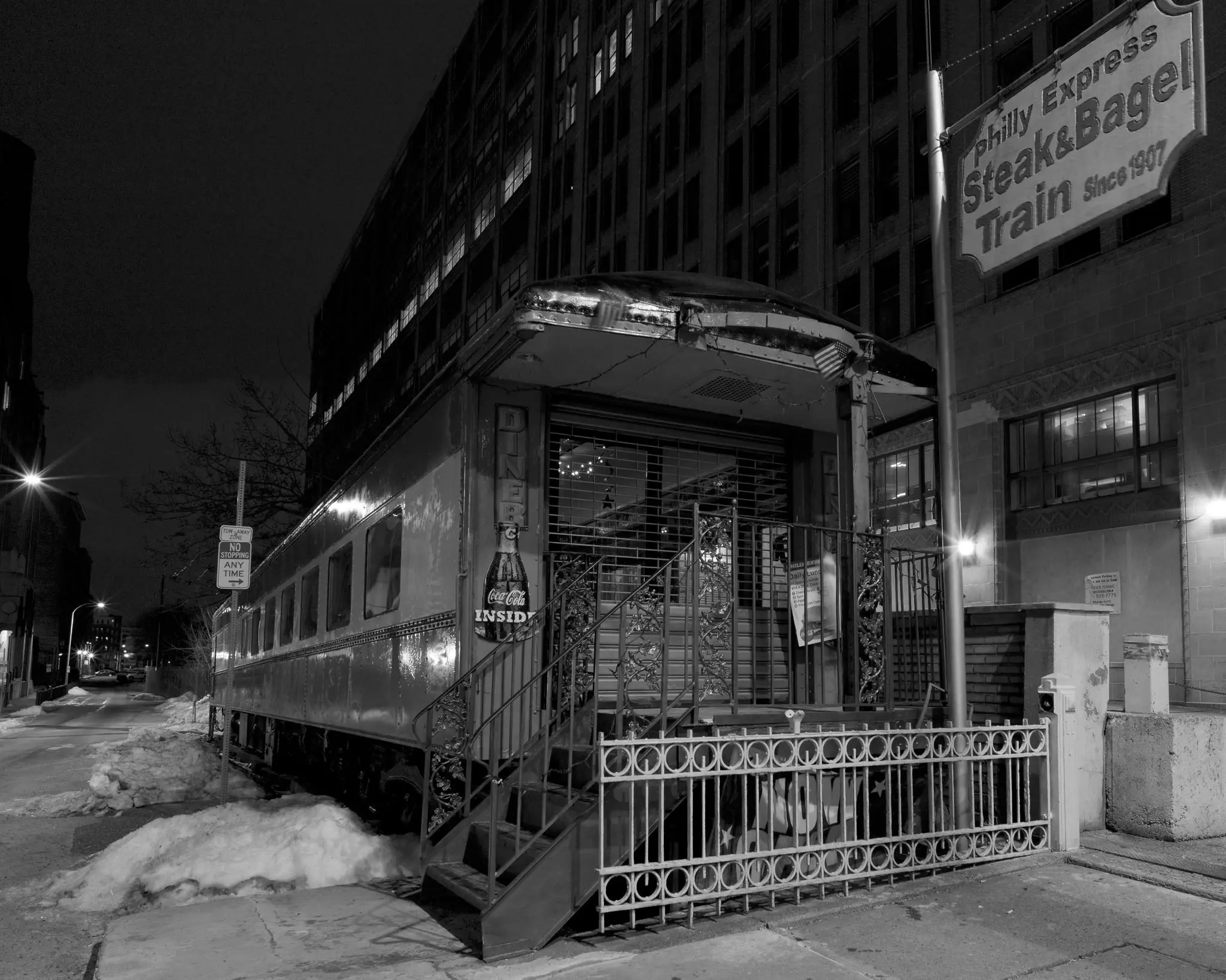 A small diner, built from a railroad car, at night, on an empty city street. A sign reads "Philly Express / Steak & Bagel Train / Since 1907"