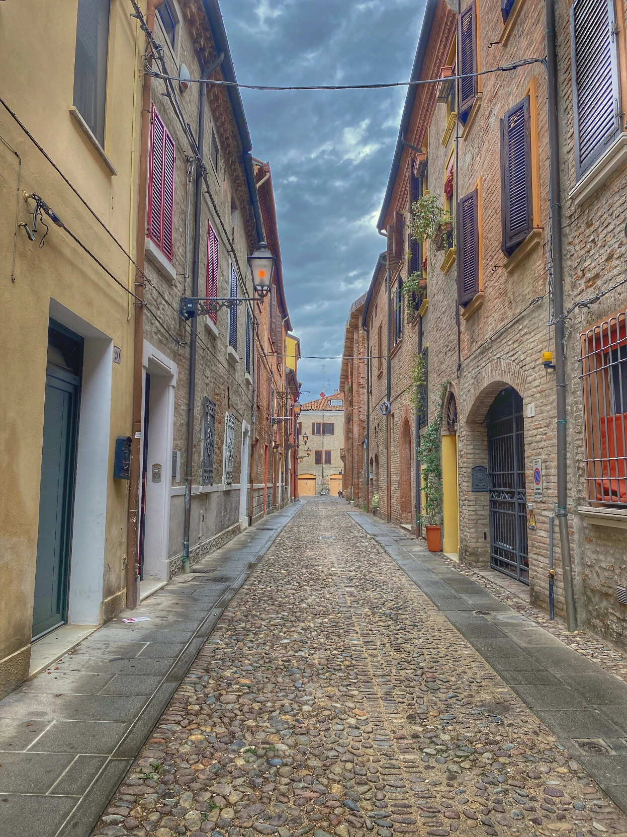 An ancient road in Ferrara, Italy. The road lies in between, on both sides a row of ancient buildings, adorned with flowers in their windows, and at the end of the street, another old edifice. The buildings are either red brick or painted in shades of yellow, while the road is paved with cobblestones. The sky, visible through a small gap left by the buildings' roofs, is cloudy. A sense of urban tranquility, of antiquity, of history, evoking introspection and memories.