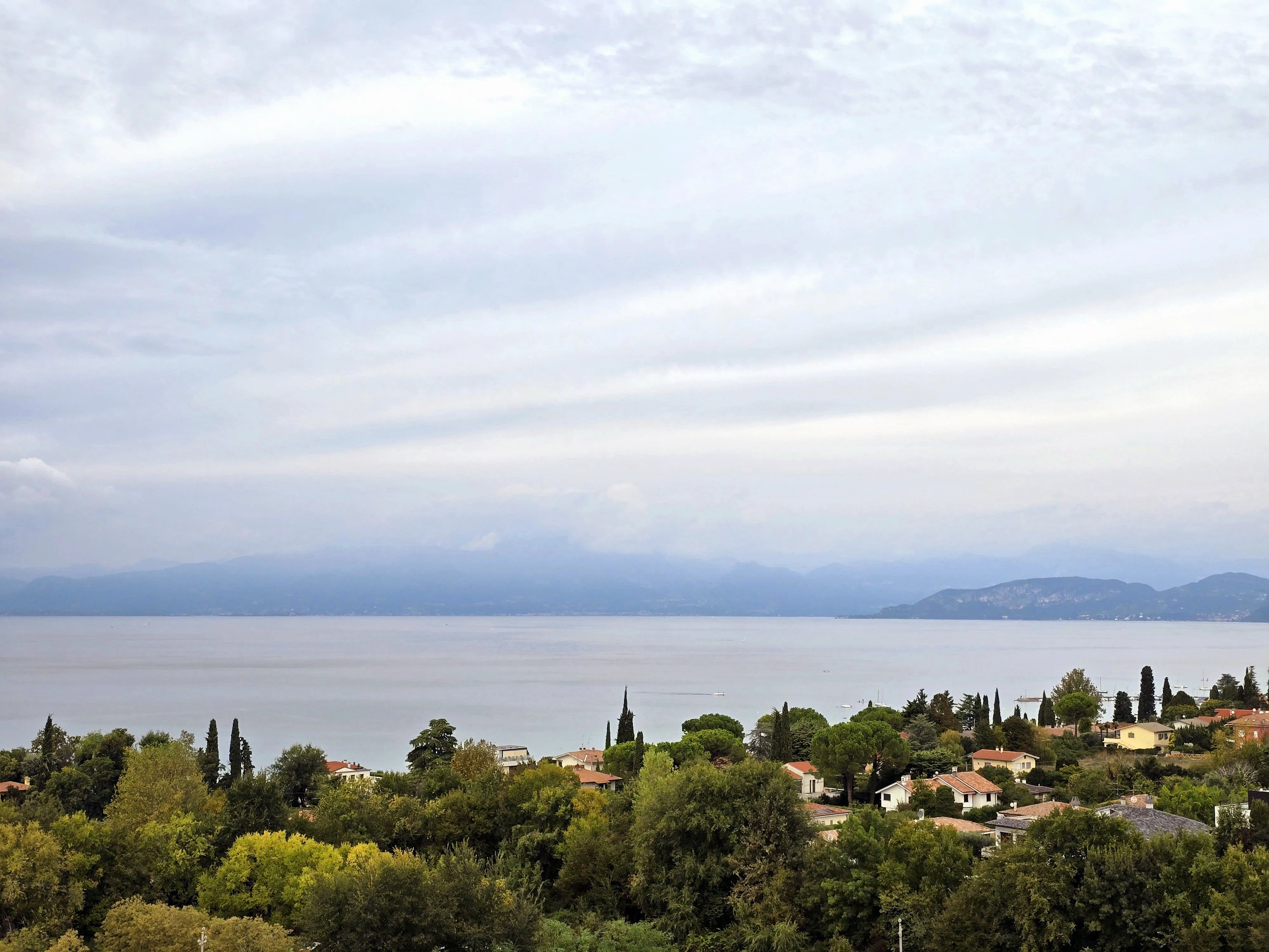 A serene view of Lake Garda, Italy, on a cloudy day. The lake stretches across the horizon, its surface calm and reflective under a soft, overcast sky. The distant mountains appear hazy, blending gently into the low-hanging clouds. Below, the lush greenery and scattered rooftops of a quaint lakeside village complete the tranquil scene, evoking a peaceful, idyllic atmosphere.