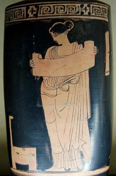 An antique greek vase, showing a woman with a scroll in her hands, looking at what appears to be a chair