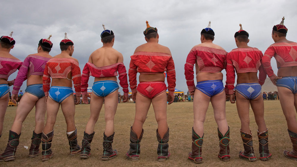 the picture shows several buff men from behind. they are wearing formal Mongolian boots, hats, bolero style shirts, and underpants with elaborate embroidery.