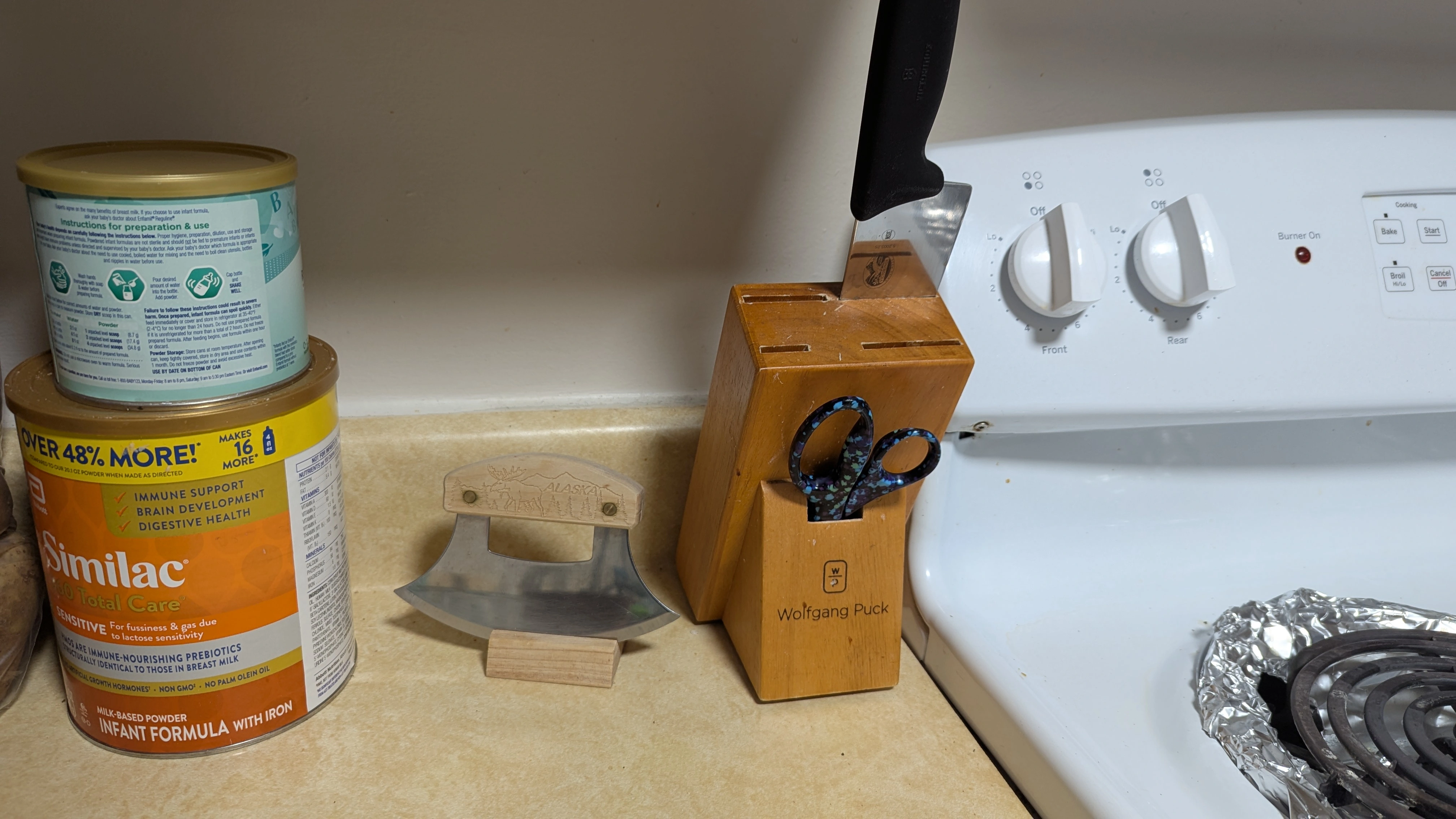 An ulu in a display block on a kitchen counter next to a garden variety knife block with 1 chefs knife in it, and a pair of scissors.